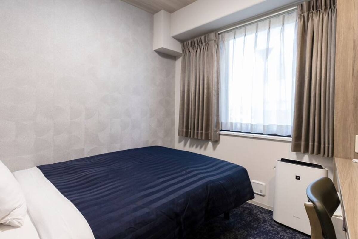 Een hotelkamer met een bed, een bureau, een stoel, een kleine verwarming en een raam met gordijnen.