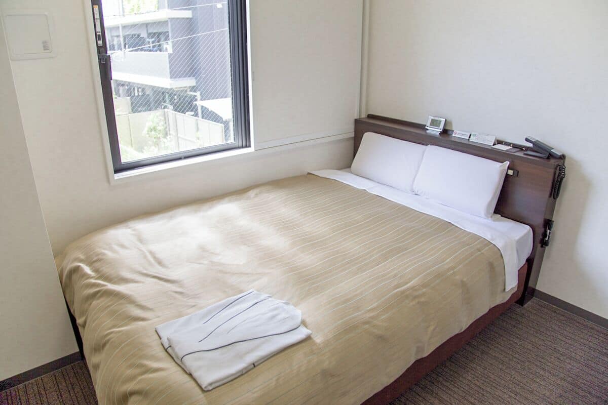 Une chambre d’hôtel avec un lit simple, une fenêtre donnant sur un paysage agréable, une table de chevet avec un téléphone et un thermostat fixé au mur.