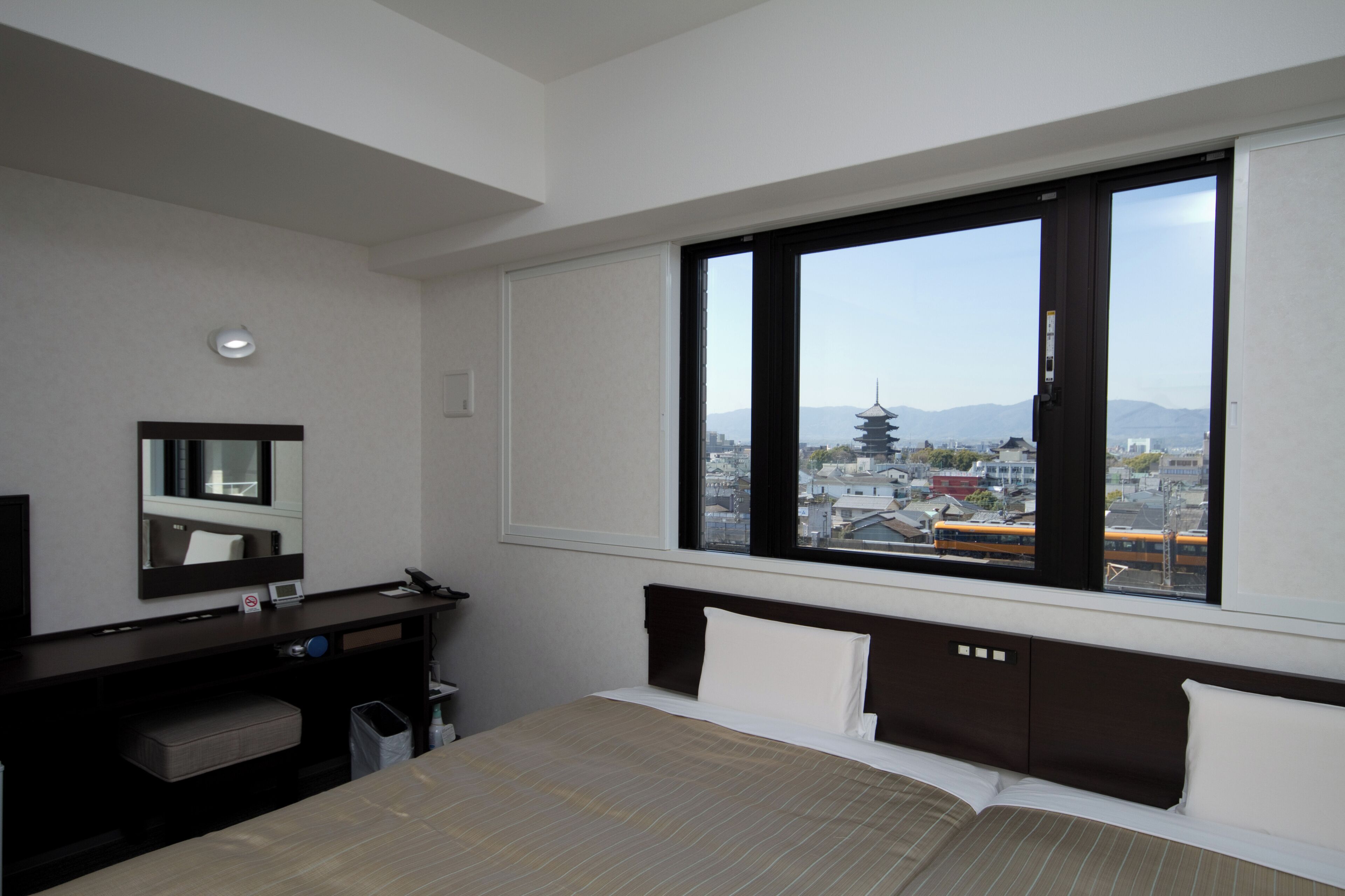 Une chambre d’hôtel équipée d’un lit, d’un bureau, d’une chaise, d’un miroir et d’une fenêtre donnant sur le paysage urbain.