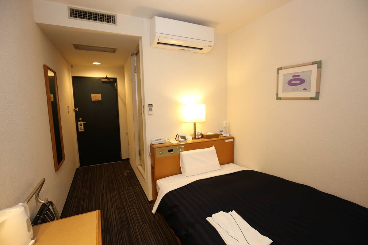 Een hotelkamer met een bed, nachtkastje, lamp en een deur naar een andere kamer.