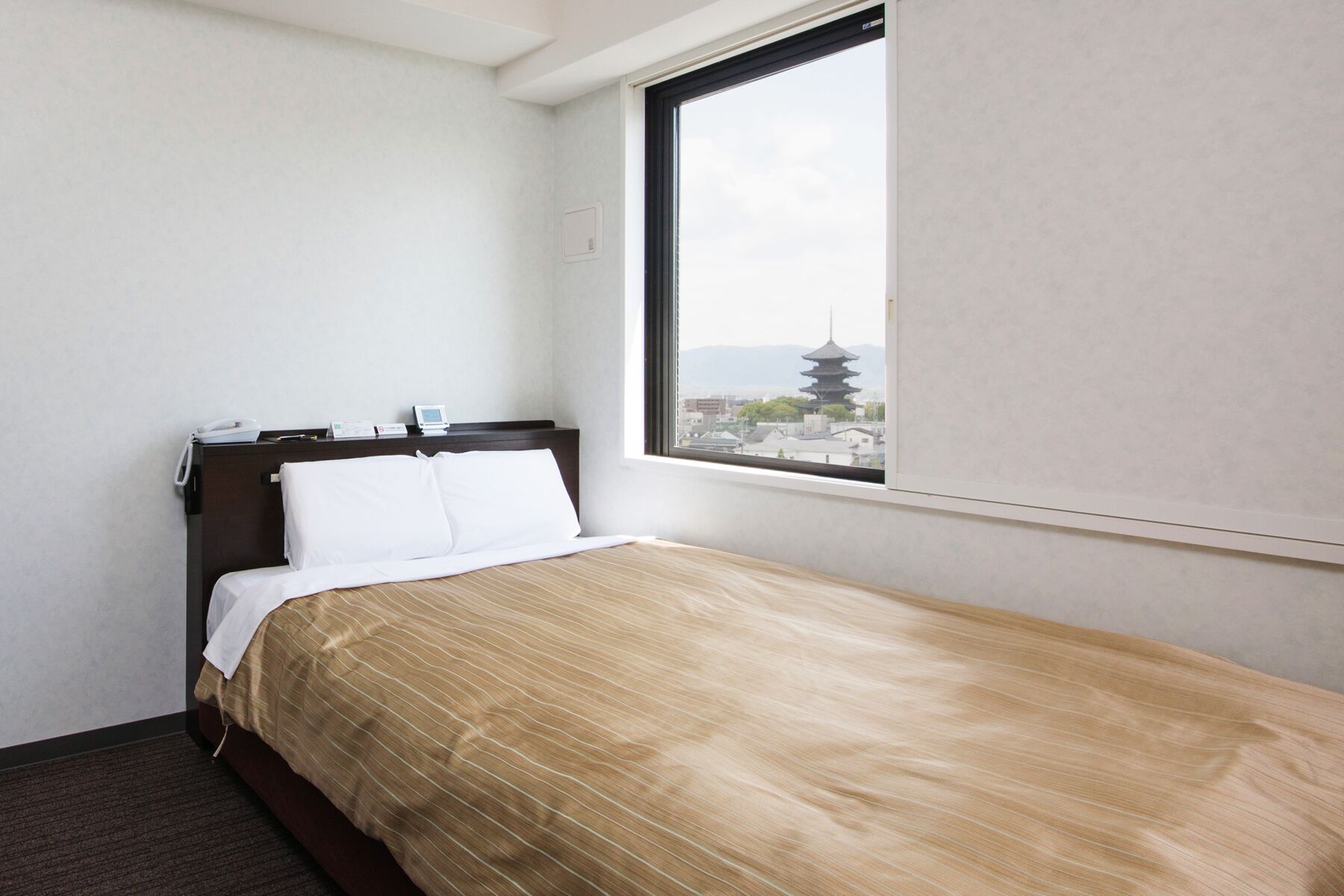 Une chambre d’hôtel avec un lit, une table de chevet sur laquelle se trouvent un téléphone et un petit appareil, une fenêtre donnant sur une pagode et un mur sobre.