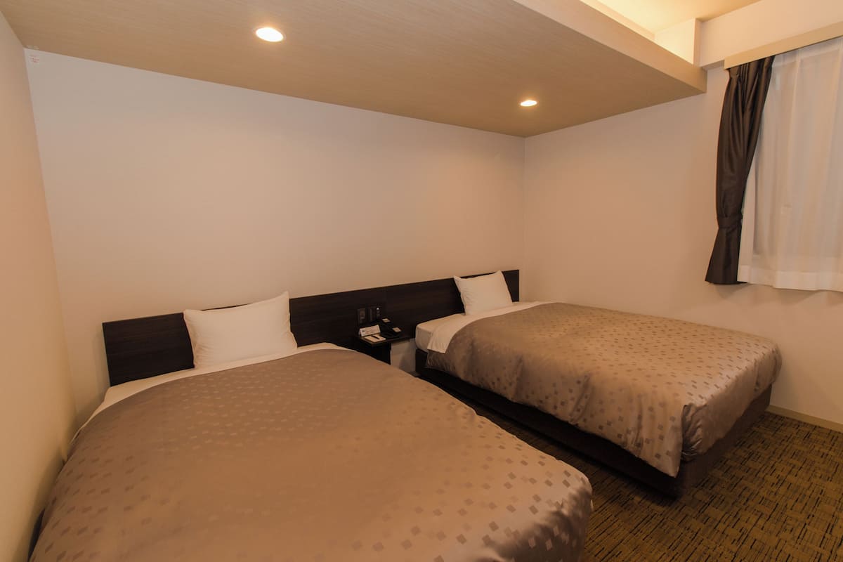 Une chambre d’hôtel avec deux lits, une table de chevet, une fenêtre avec des rideaux et un luminaire au plafond.