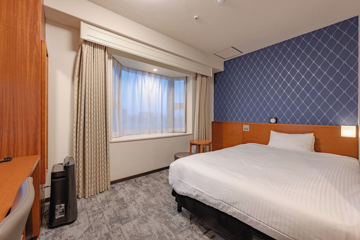 A hotel room with a bed, a desk, a chair, a waste bin, and a window with curtains.
