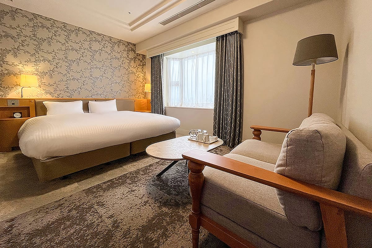 A hotel room with a large bed, a sofa, a small table, a lamp, and a window with curtains.