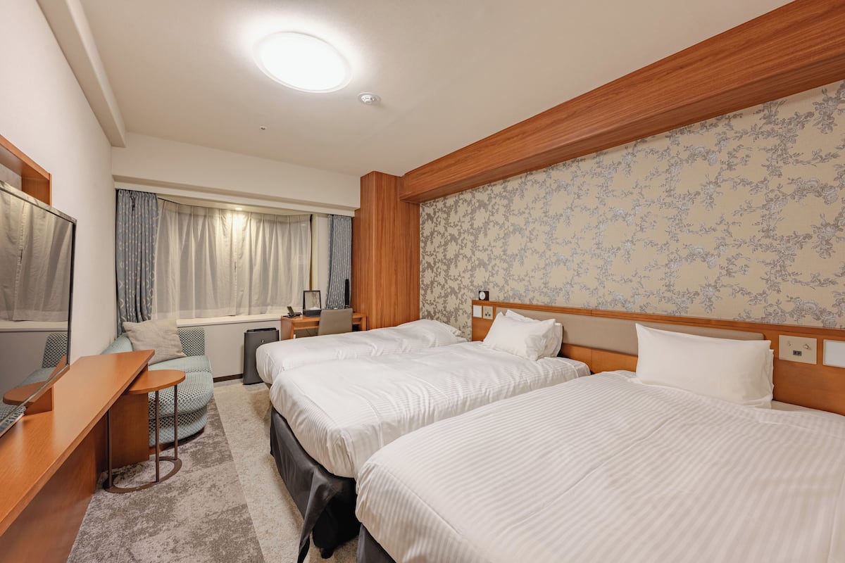 A hotel room with two beds, a desk, a chair, a small table, a window with curtains, and a floral wallpaper.