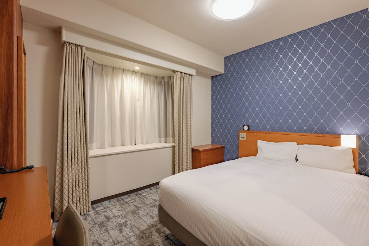 A hotel room with a large bed, a wooden nightstand, a desk with a chair, and a window with curtains.