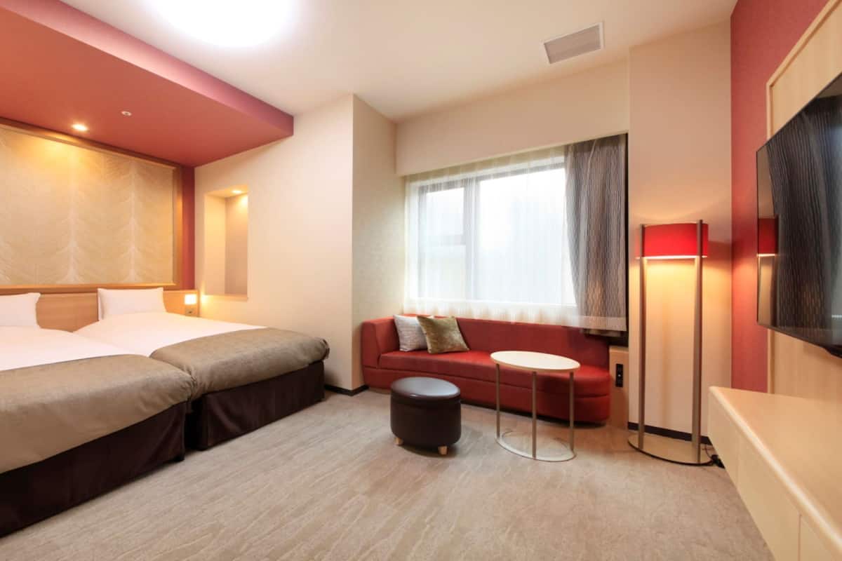 A hotel room with two beds, a sofa, a small table, a TV, and a window with curtains.