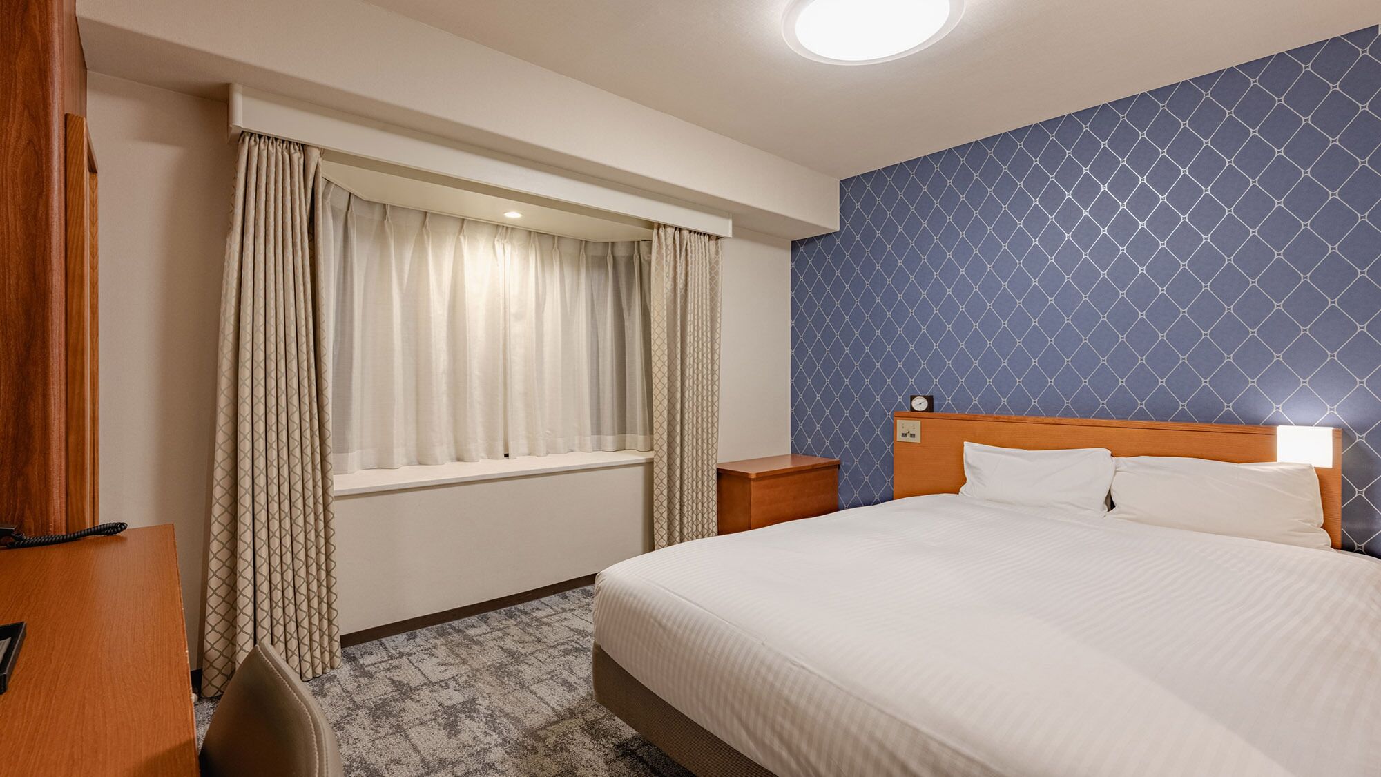 Hotellihuone, jossa on sänky, yöpöytä, kirjoituspöytä puhelimella ja ikkuna, jossa on verhot.