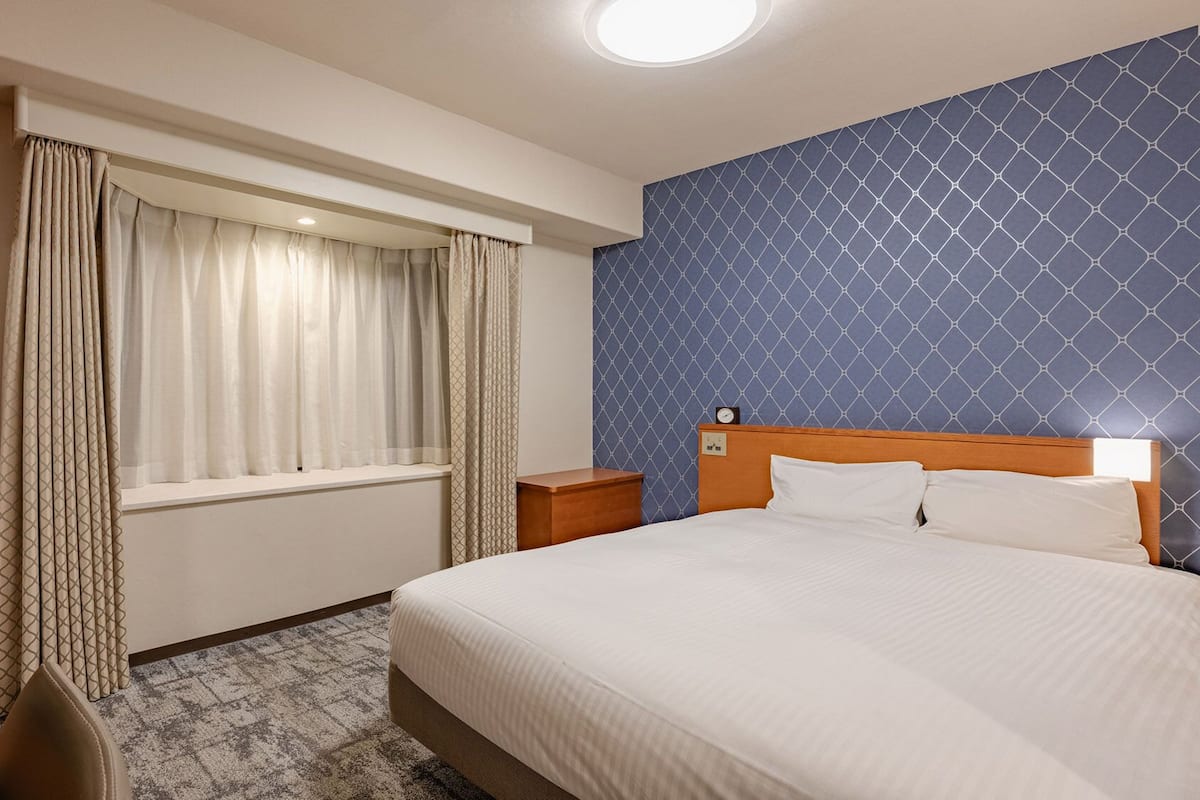 A hotel room with a bed, a nightstand, a desk with a telephone, and a window with curtains.