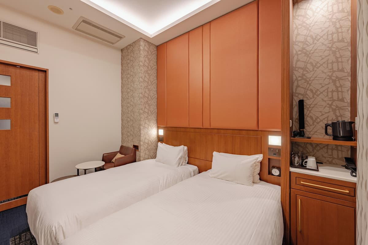 A hotel room with two beds, a small table, a chair, a wardrobe, and a shelf with a kettle and a coffee maker.