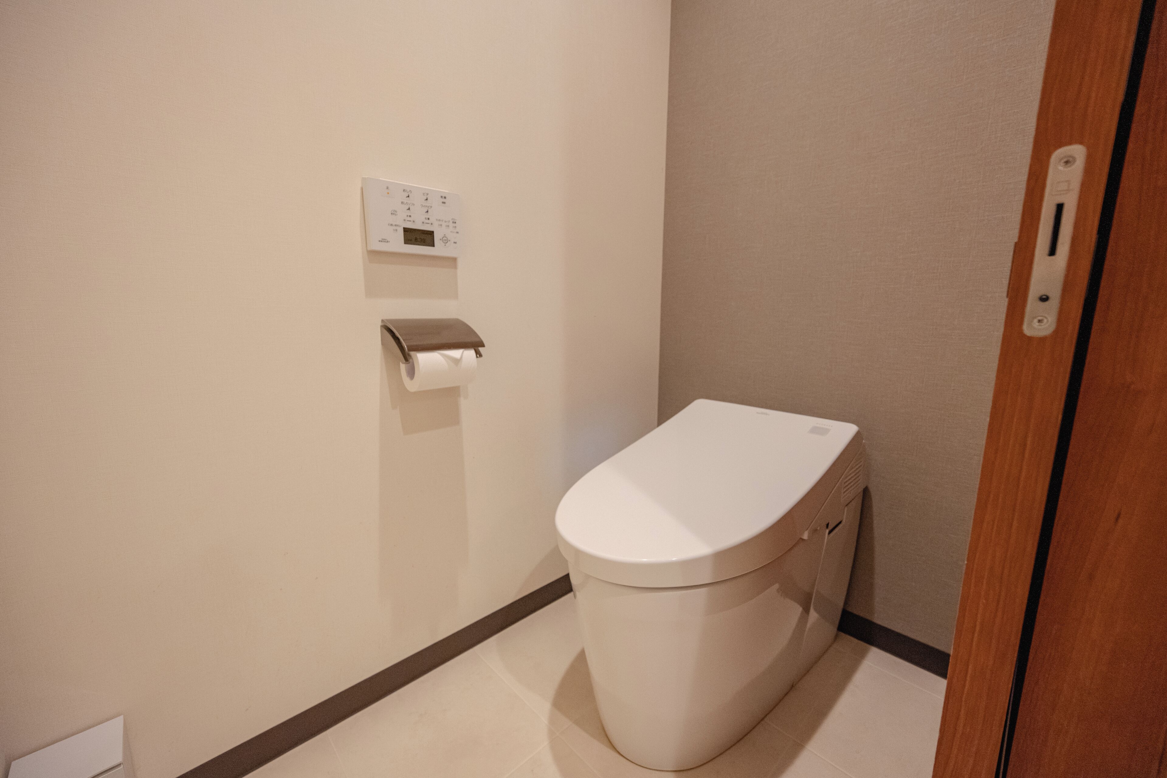 A small, clean bathroom with a toilet, a paper towel dispenser, and a wall-mounted control panel.