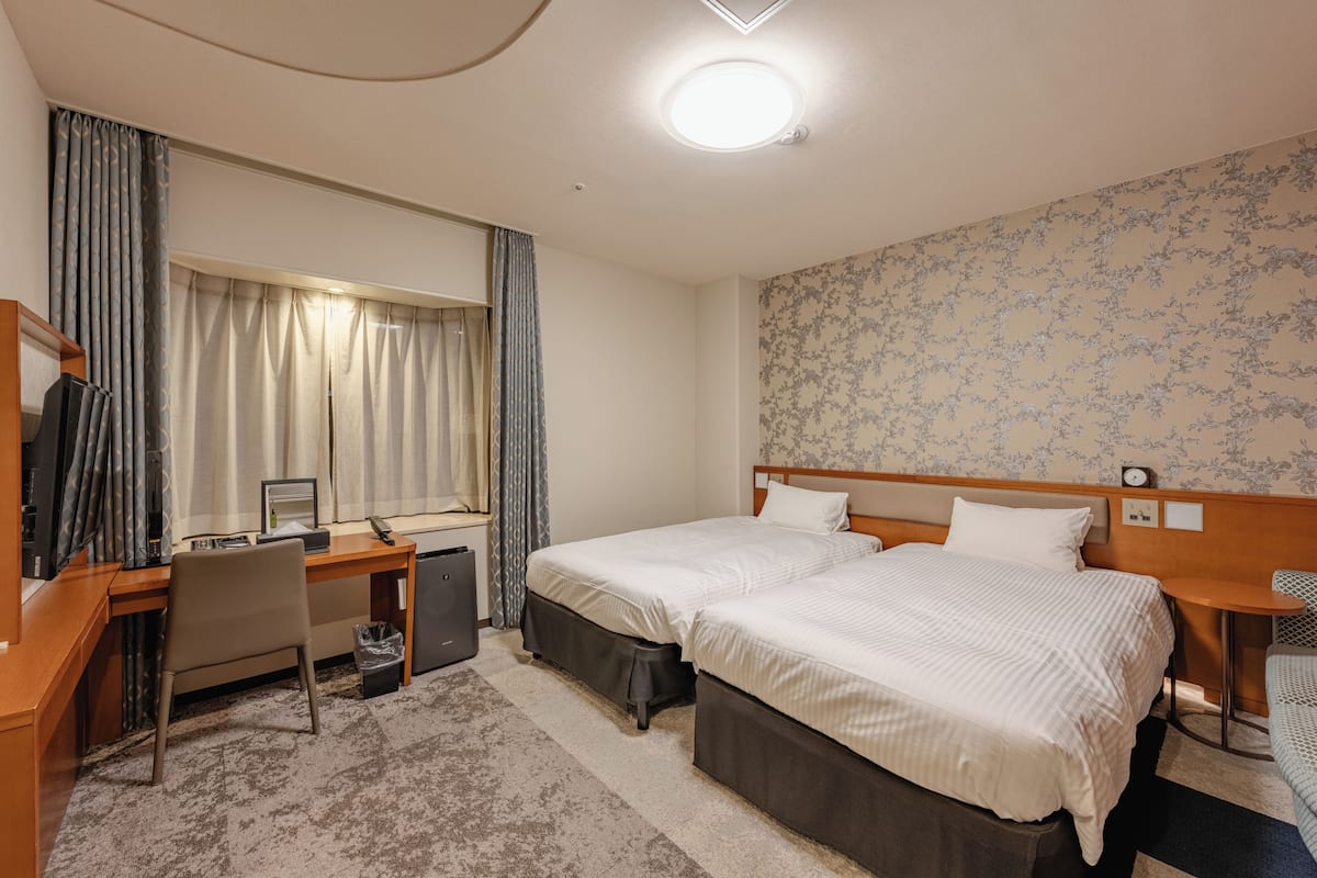 A hotel room with two beds, a desk with a computer, a chair, a small table, and a window with curtains.