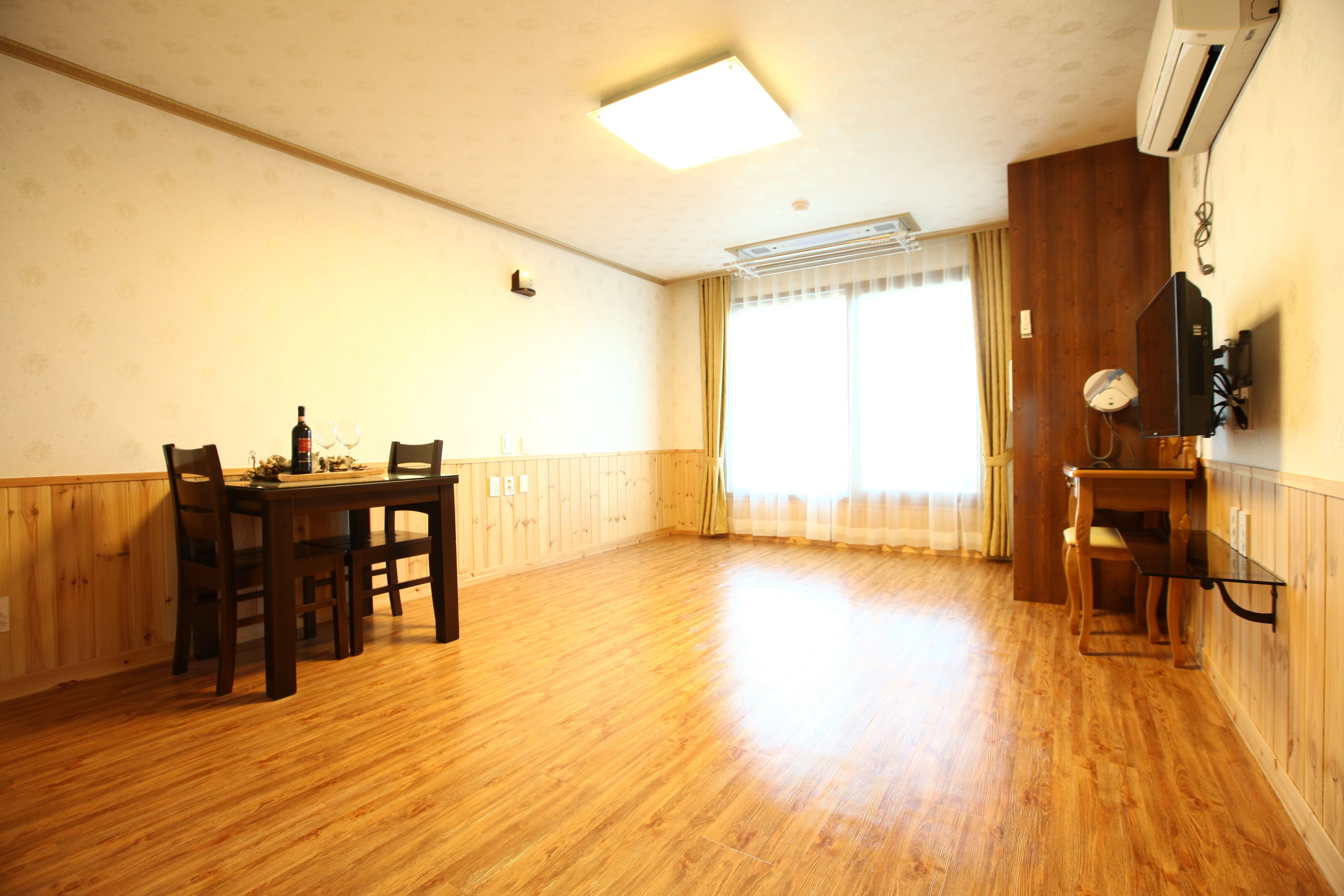 A room with wooden flooring, a dining table with chairs, a TV mounted on the wall, and a window with curtains.