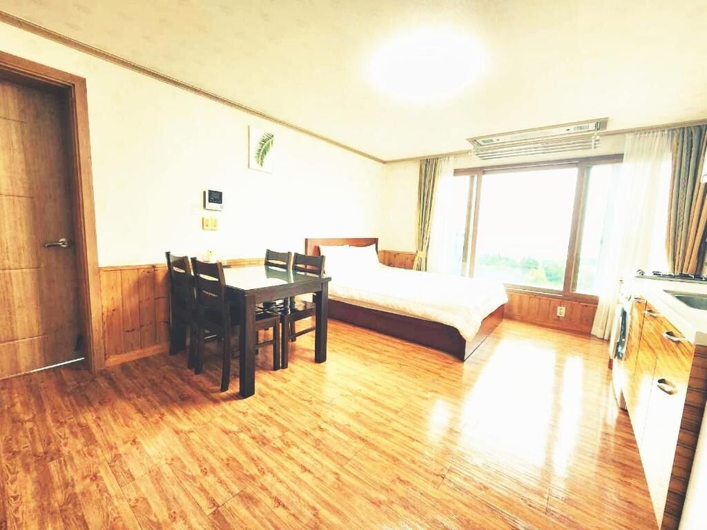 A hotel room with a wooden floor, a bed with white bedding, a desk with chairs, a window with curtains, and a kitchenette area.