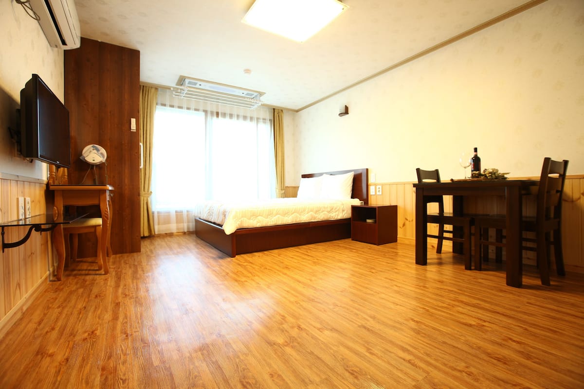 A hotel room with a wooden floor, a bed with white bedding, a television mounted on the wall, a desk with chairs, and a window with curtains.