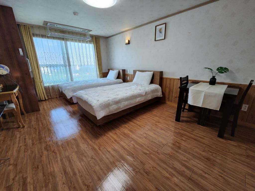 A room with two beds, a wooden floor, a desk with a chair, and a window with curtains.