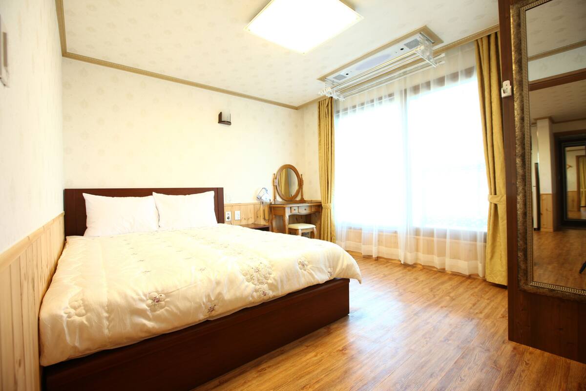 A hotel room with a large bed, wooden flooring, a bedside table with a mirror, and a window with curtains.