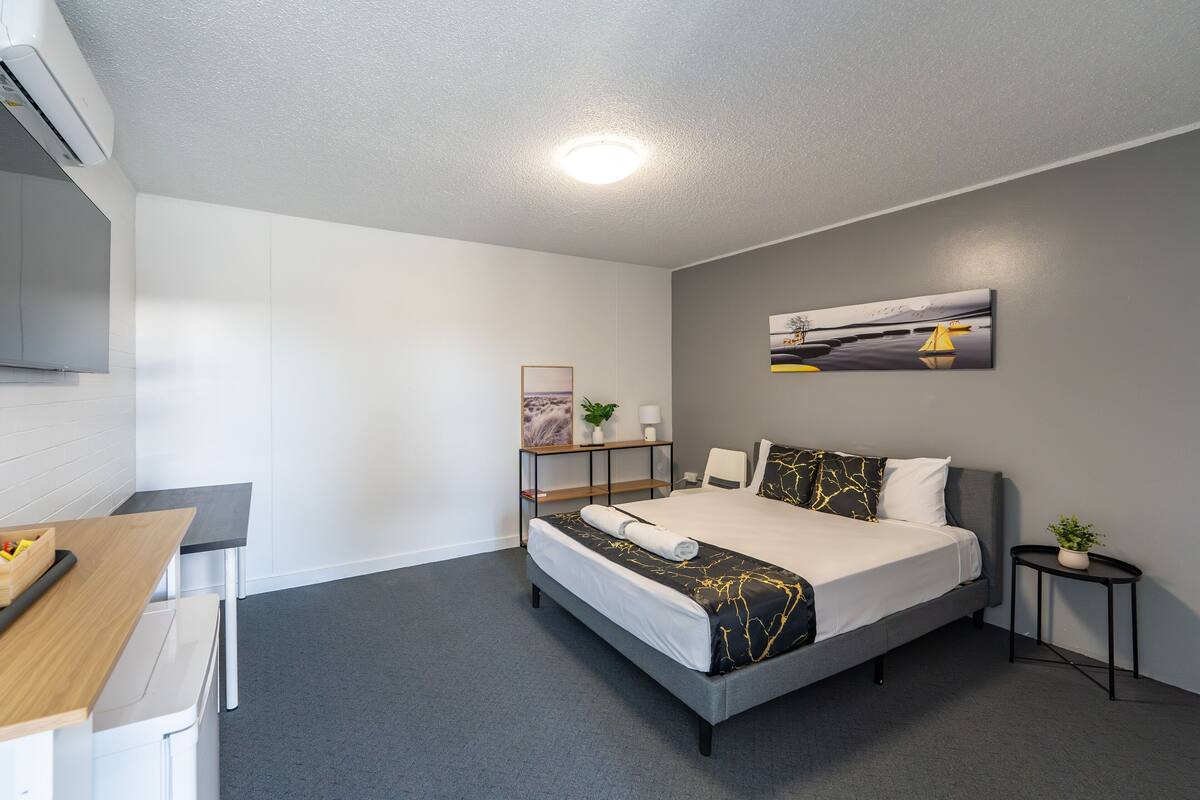 A modern hotel room with a grey carpet, a bed with a black and gold bedspread, a small bedside table with a plant, and a painting on the wall.