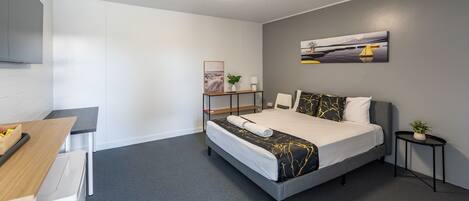 Una habitación de hotel moderna con alfombra gris, una cama con colcha negra y dorada, una mesita de noche con una planta y un cuadro en la pared.