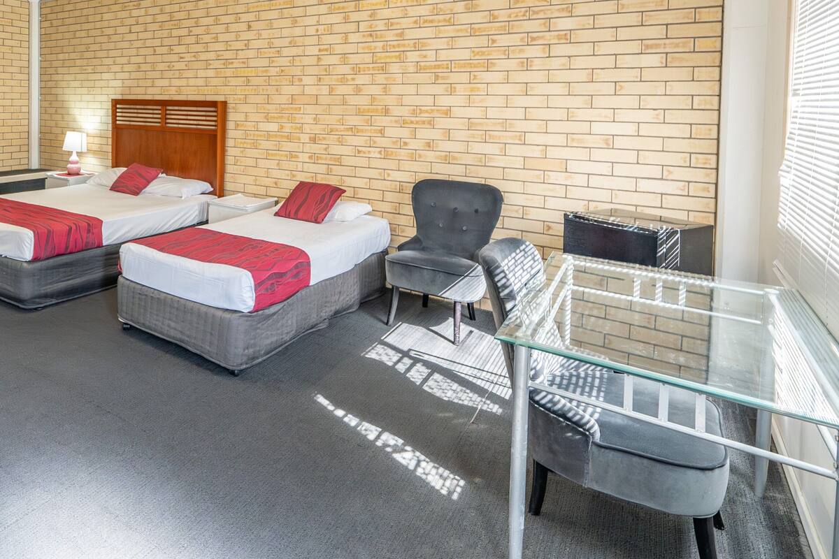 A hotel room with two beds, a chair, a glass table, and a brick wall.
