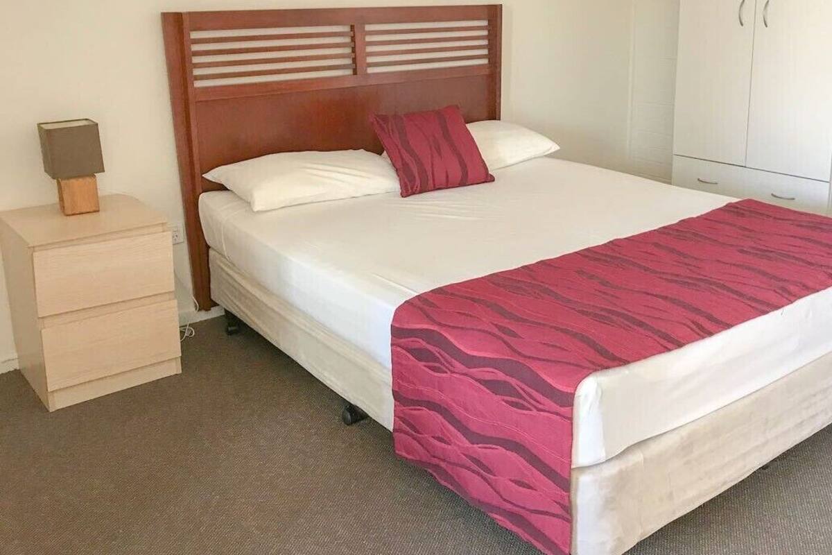 A neatly made bed with a wooden headboard, a bedside table with a lamp, and a wardrobe in a hotel room.