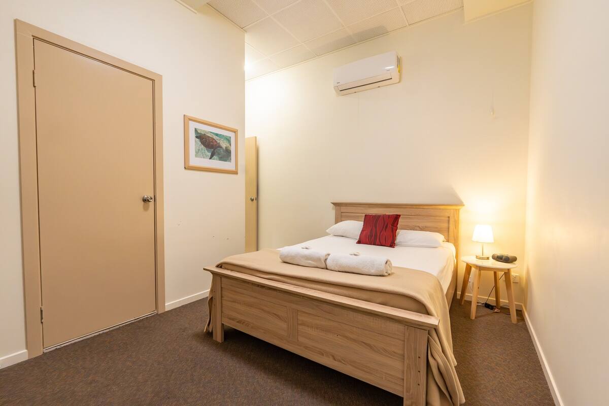 A hotel room with a wooden bed, a bedside lamp, an air conditioner, a door, and a framed picture on the wall.
