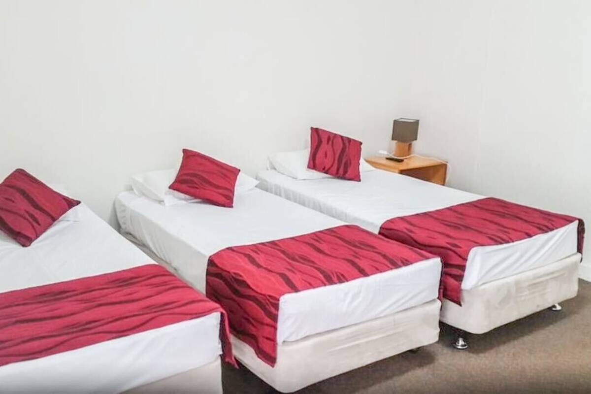 A hotel room with four single beds, each with a white mattress and red bedding. There is a small wooden nightstand with a lamp and a phone.