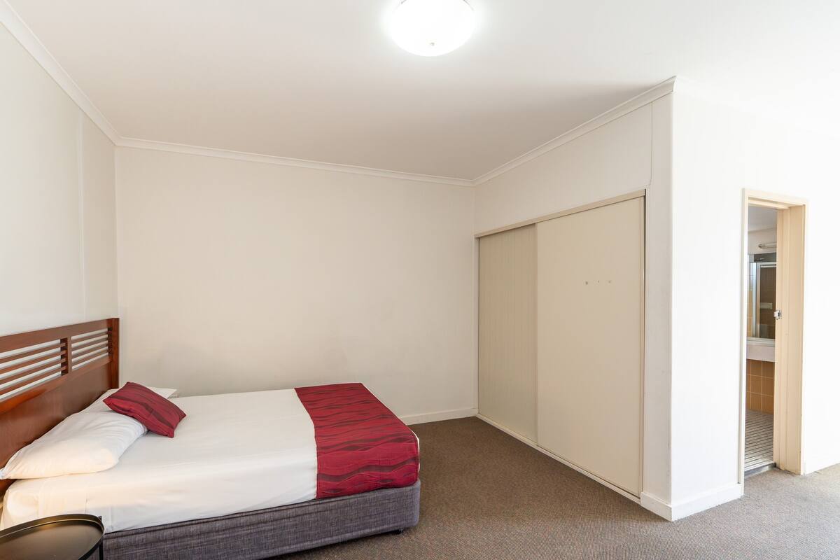 A single bed with a wooden headboard, a red and white bedspread, and a small bedside table with a lamp.