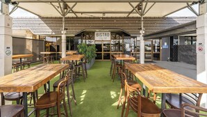 Un bistro al aire libre con mesas y sillas de madera, piso de césped artificial verde y estructura de techo con tejas.
