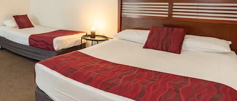 A hotel room with two beds, each with a wooden headboard, white bedding, and red decorative pillows. There is a small bedside table with a lamp between the beds.
