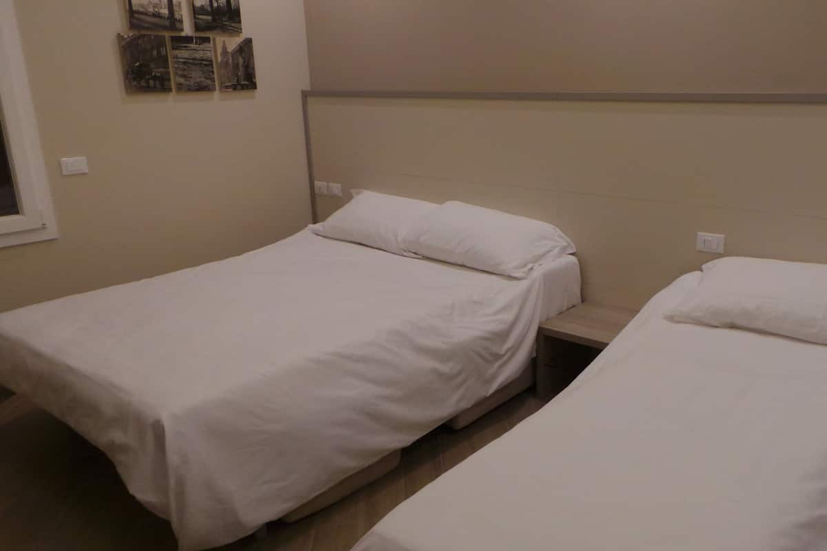 Habitación con dos camas individuales, cada una con ropa de cama blanca. Se observan interruptores de luz en la pared y una ventana pequeña a la izquierda.