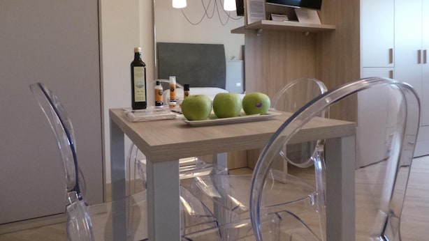 A modern hotel room with a wooden table, clear chairs, a TV on a shelf, and a mirror. - Marbia Guest House (Bologna)
