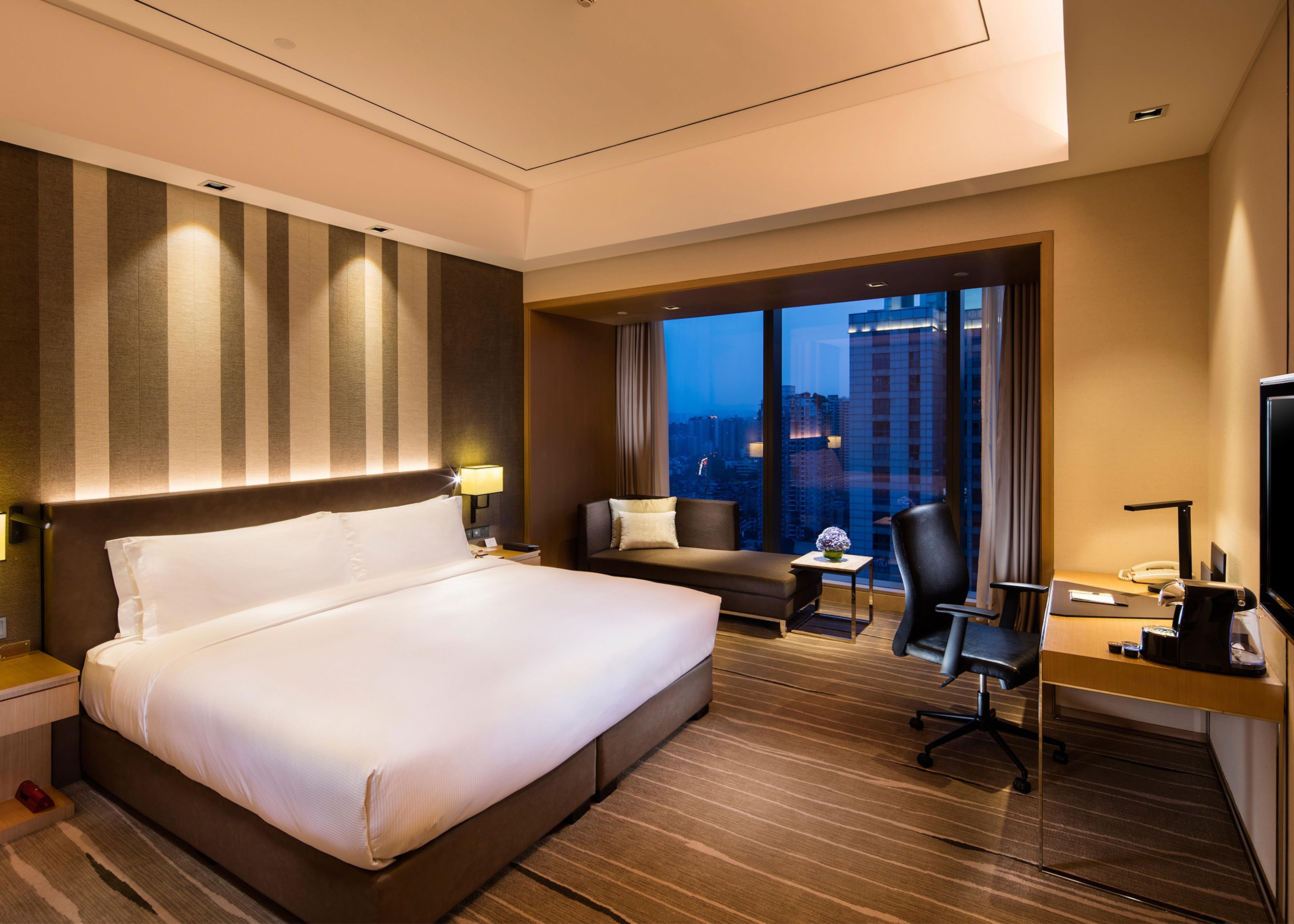 A hotel room with a large bed, a desk with a chair, a sofa, and a view of the cityscape.