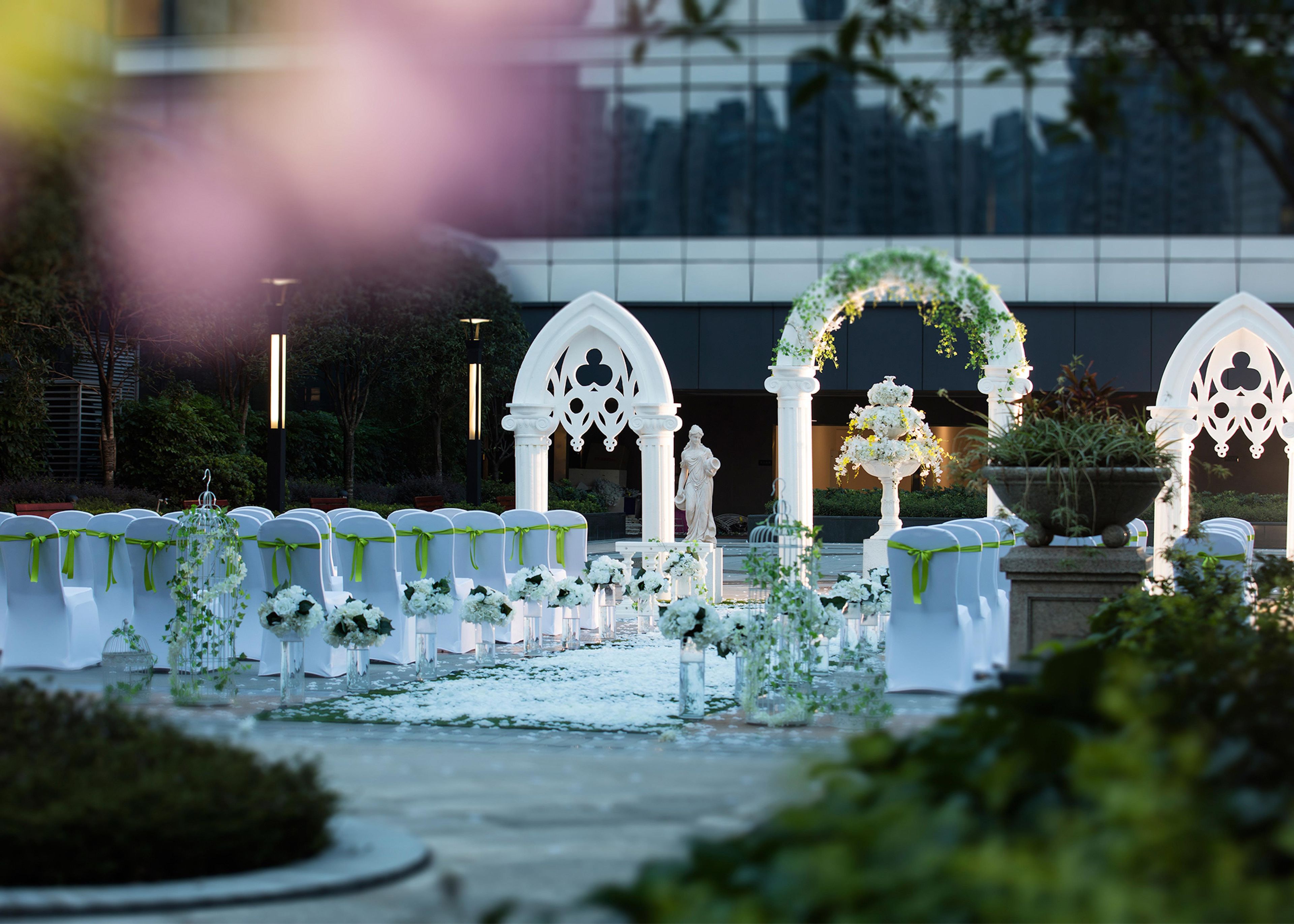 Un área de estar al aire libre con ambiente formal, con sillas y mesas blancas, decorada con arreglos florales y follaje.