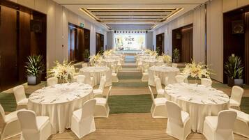 A banquet hall with round tables set for dining, white chairs, and potted plants.