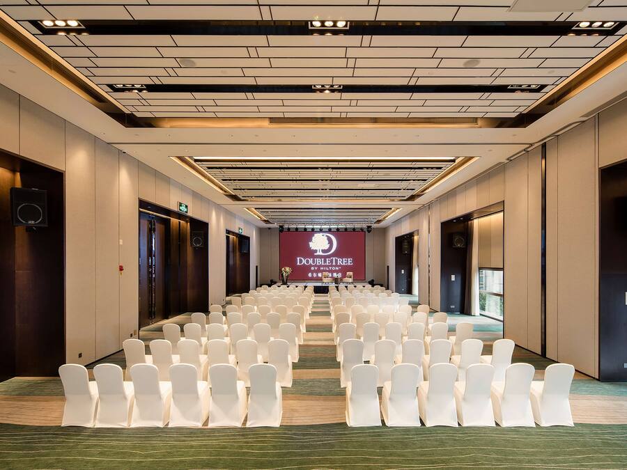 Een vergaderzaal met rijen witte stoelen, een groot scherm waarop 'DubleTree by Hilton' staat, en een modern plafondontwerp.