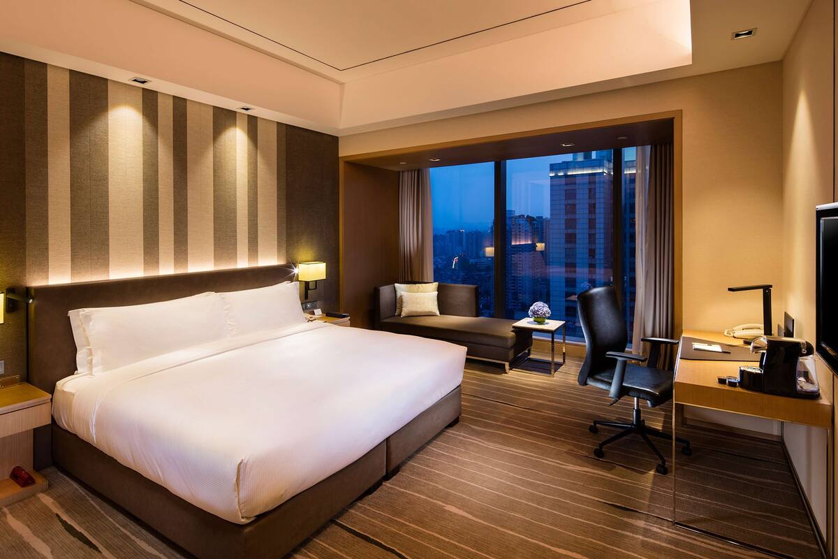 Une chambre d’hôtel avec un grand lit, un bureau avec un ordinateur, une chaise et une vue sur la ville la nuit.