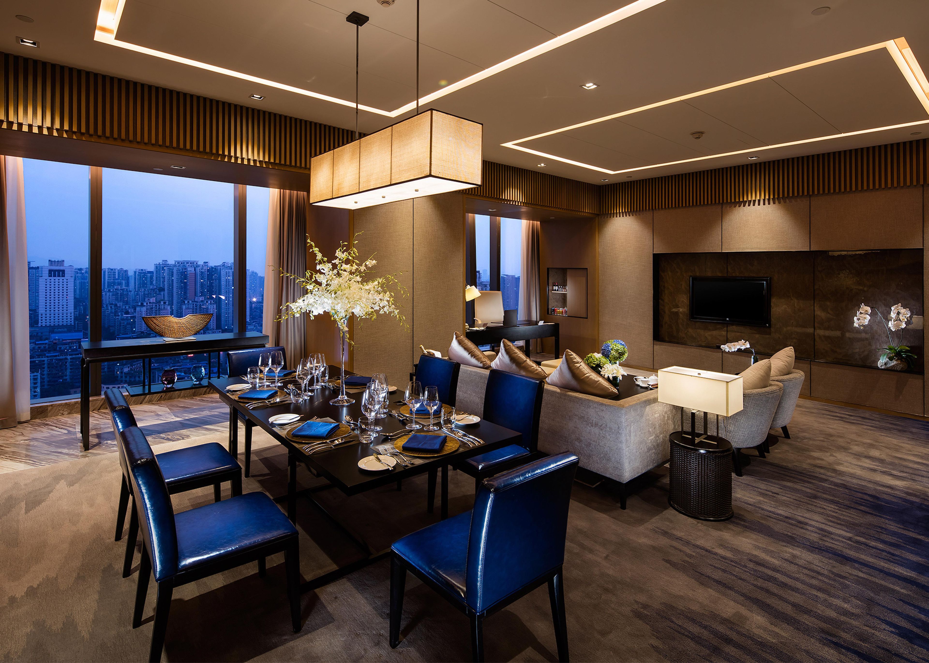 A modern hotel room with a dining area, seating, a large window with a city view, and a flat-screen TV.