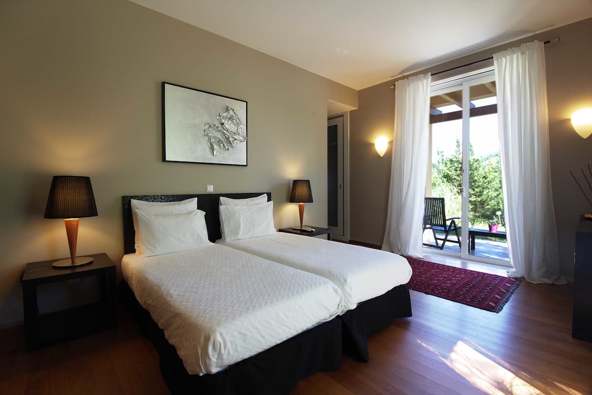 Una cama doble con ropa blanca, dos mesitas de noche con lámparas, una obra de arte enmarcada sobre la cama y vistas al exterior a través de un ventanal corredero.