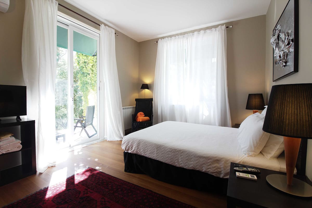 Un dormitorio con cama, dos lámparas de noche, televisor, alfombra roja y vista al exterior a través de una puerta corredera.