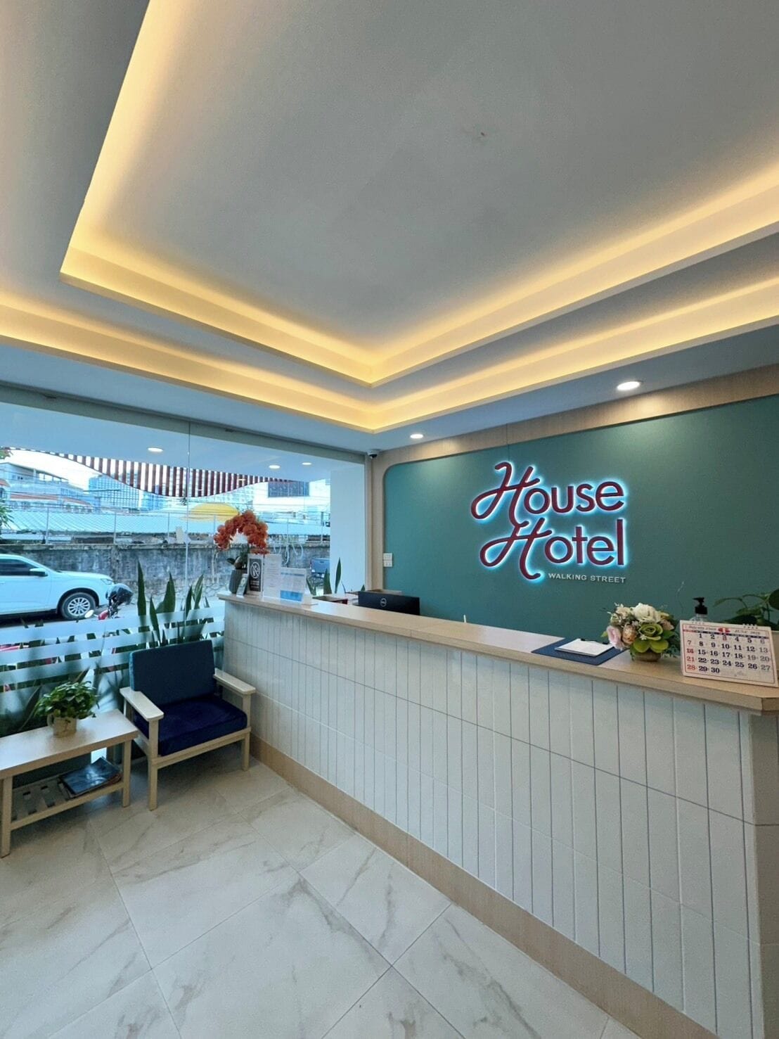 A hotel reception area with a neon sign, a reception desk, a chair, a calendar, and a view of the street outside.