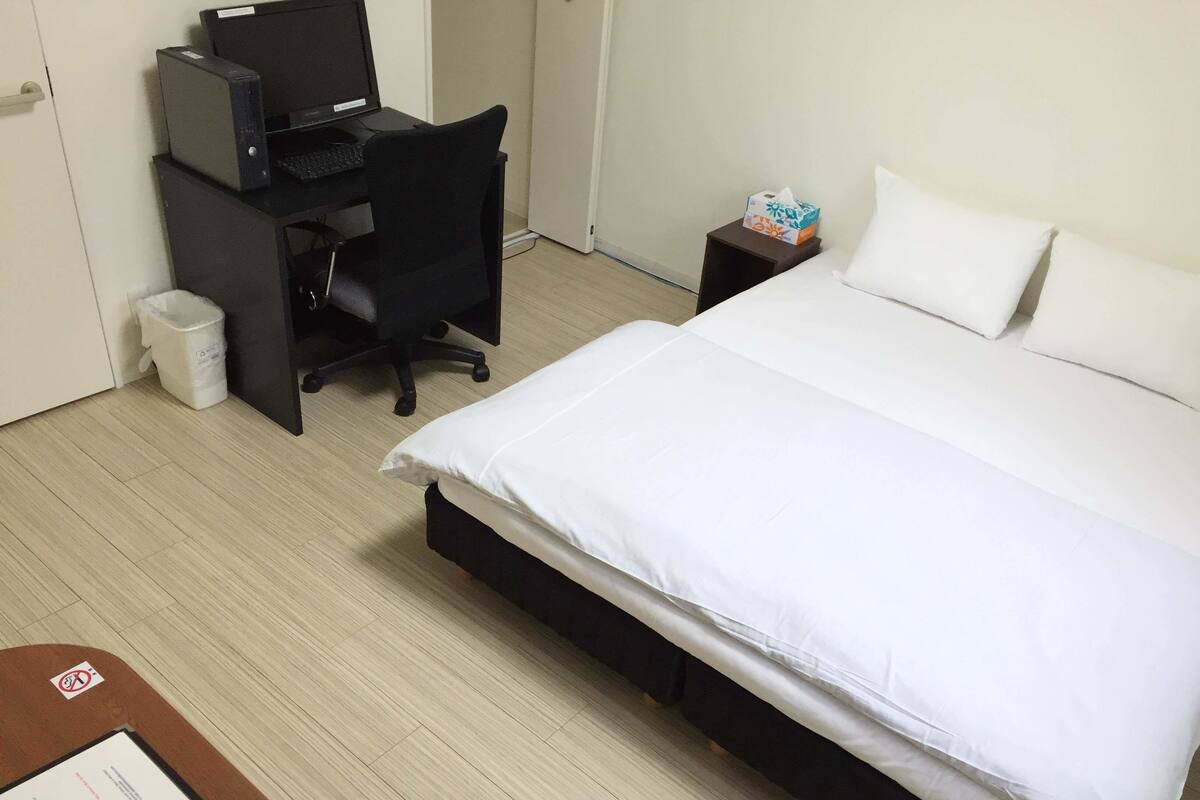 Een hotelkamer met een bed, een bureau met een computer, een stoel en een kleine prullenbak.