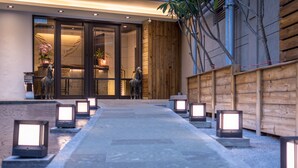Un chemin bordé de lanternes illuminées mène à l’entrée d’un bâtiment dont la façade est en bois et la porte en verre.