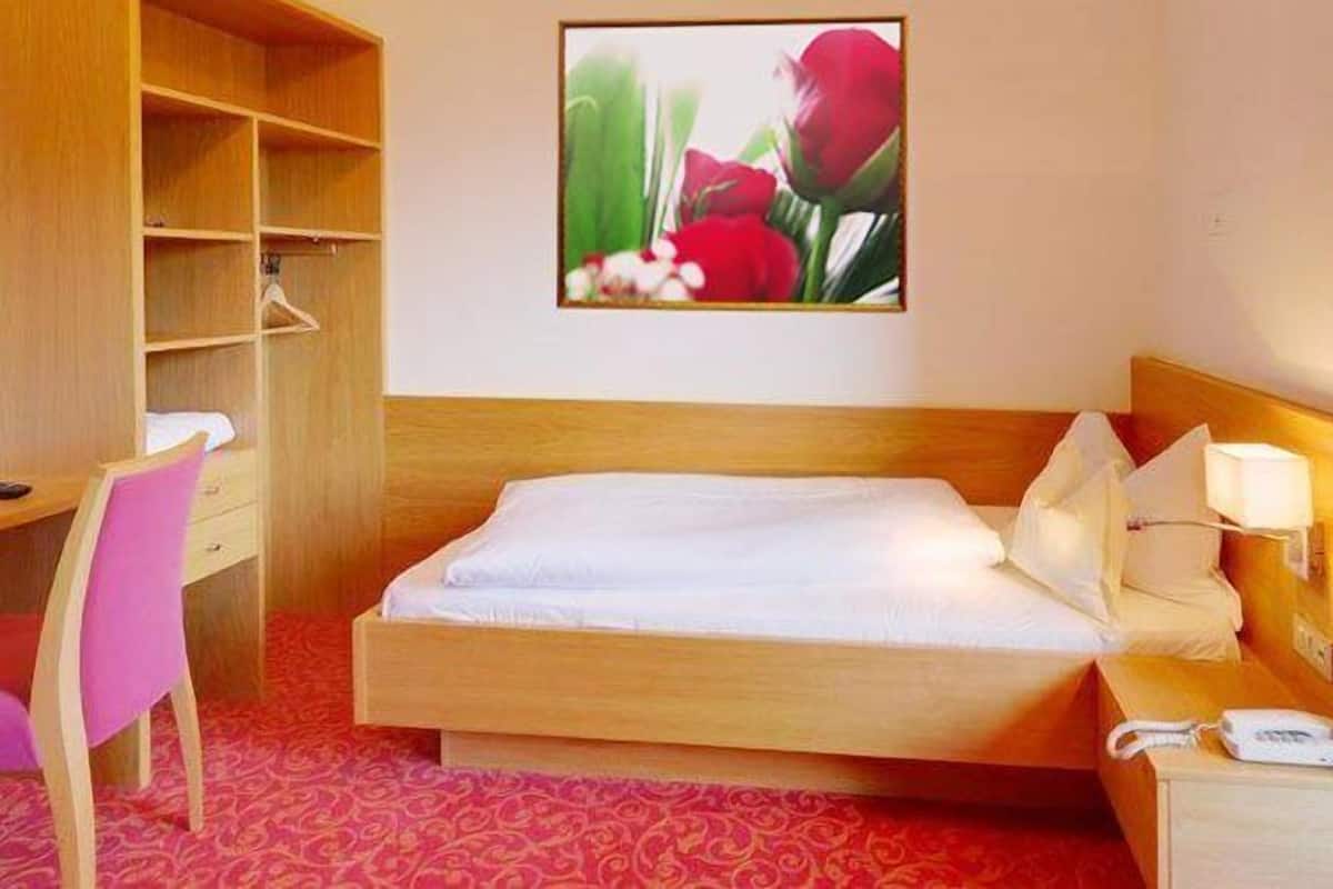 Une chambre d’hôtel comprenant un lit, une table de chevet avec une lampe, un bureau avec une chaise, une télévision et une étagère fixée au mur.