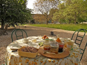 An outdoor dining table set with food and drinks, surrounded by chairs, in a garden area with trees and a building in the background. - La Ferme des Isles (Autheuil-Authouillet)