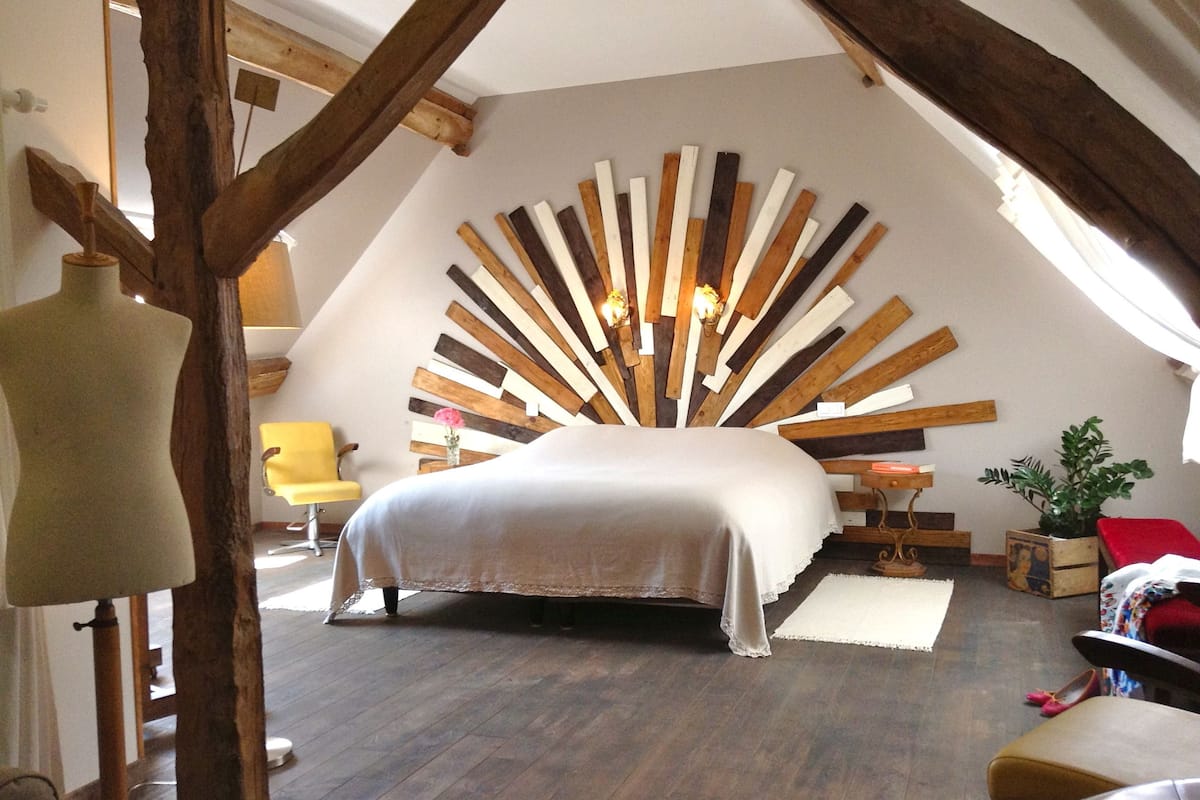 A bedroom with a sloped ceiling, a bed with a white bedspread, a wooden headboard, a yellow chair, a mannequin, and a small table with a vase of flowers.