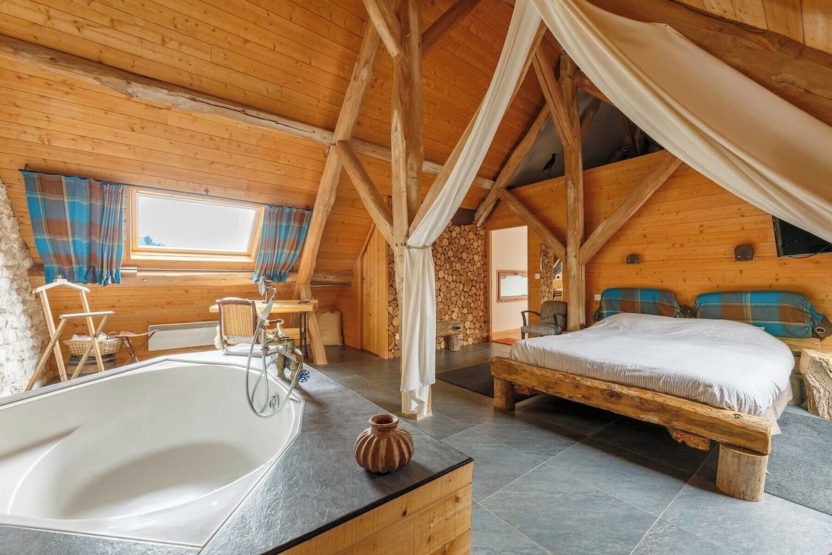 A rustic bedroom with a large wooden bed, a separate bathroom with a bathtub, and a window with plaid curtains.