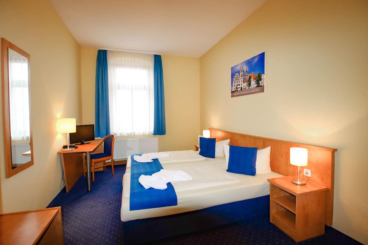 Ein Hotelzimmer mit zwei Betten, einem Schreibtisch, einem Stuhl, einer Lampe und einem Fenster mit Vorhängen.