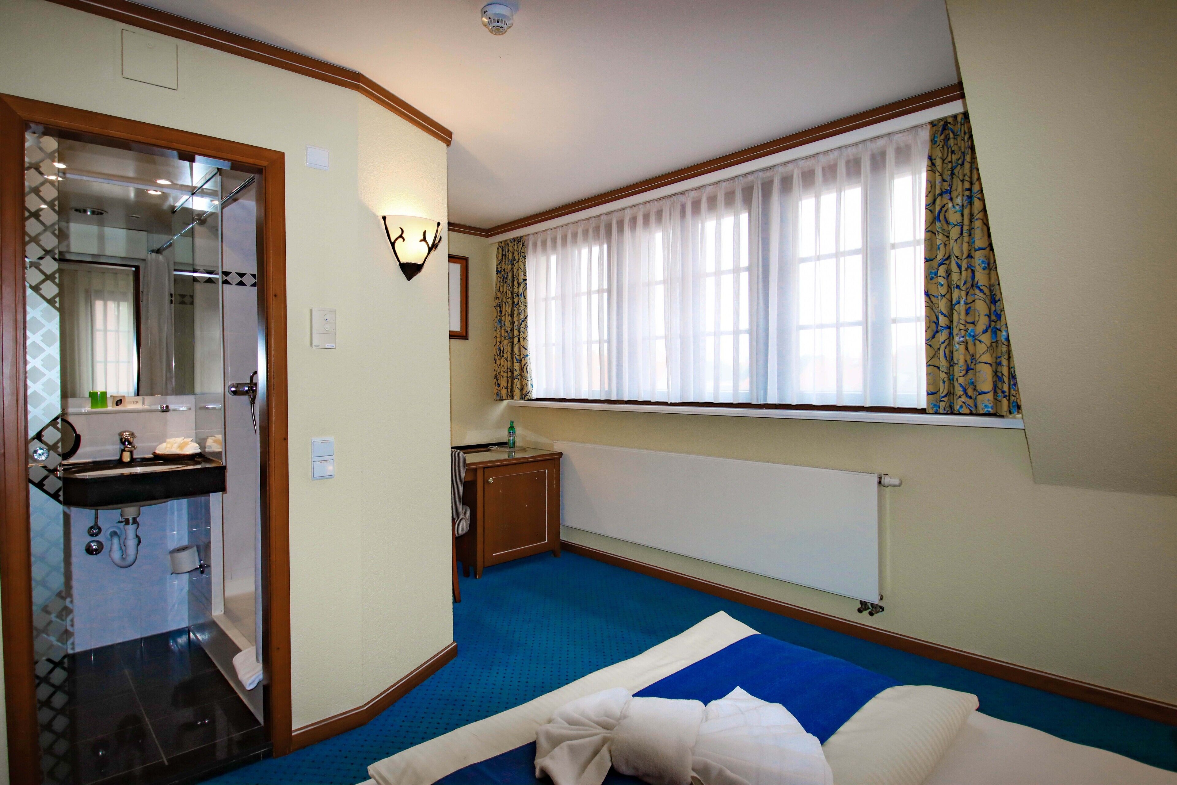 A hotel room with a bathroom, a bed, a window with curtains, and a radiator.