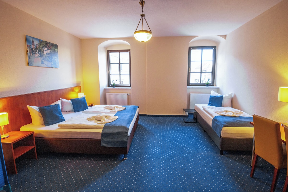 Ein Hotelzimmer mit zwei Betten, einem Schreibtisch und einem Stuhl. Es gibt zwei Fenster und ein Bild an der Wand.
